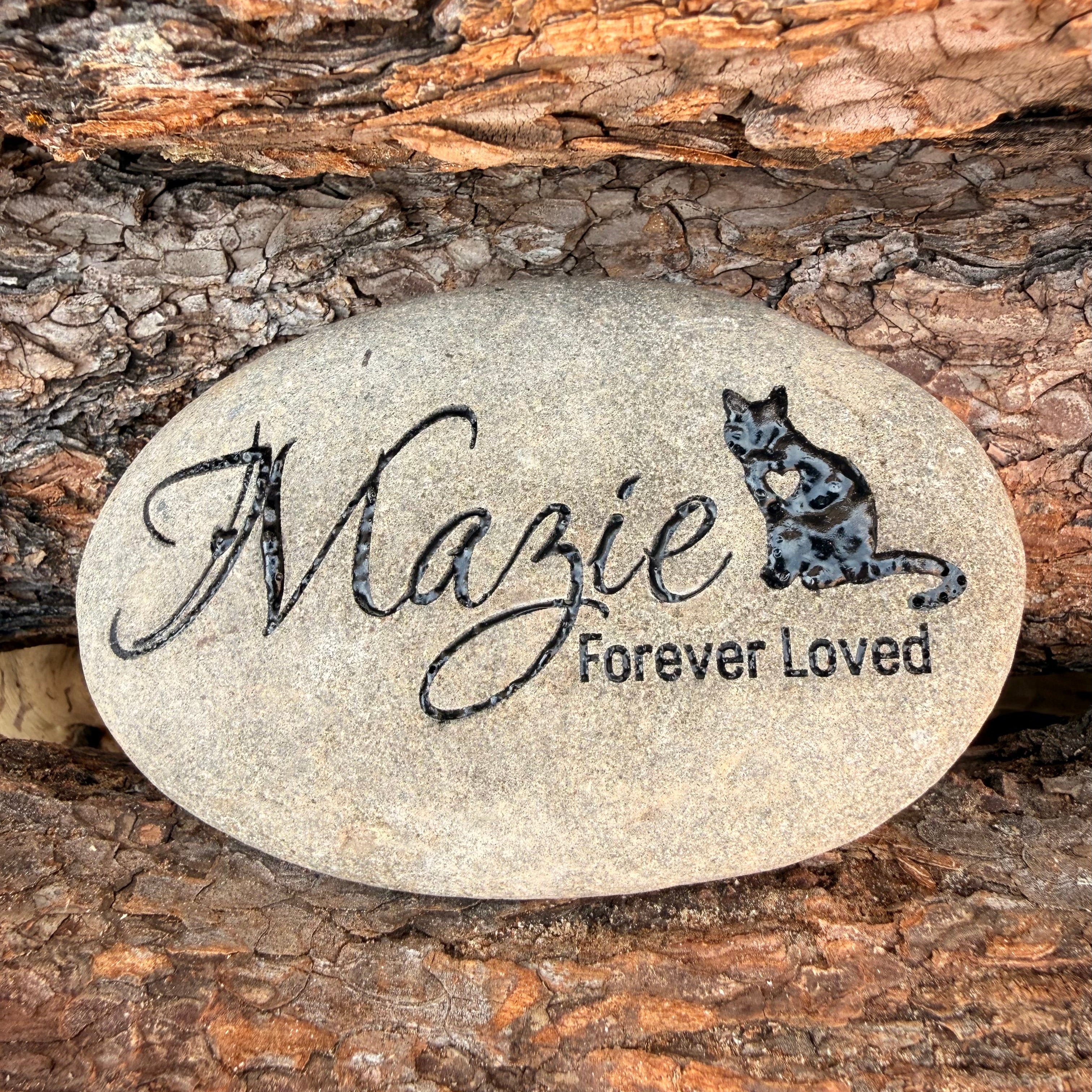 Oval-shaped stone with memorial engraving of pet's name 'Mazie', memorial statement 'Forever Loved' and a cat illustration against a wooden bark background.