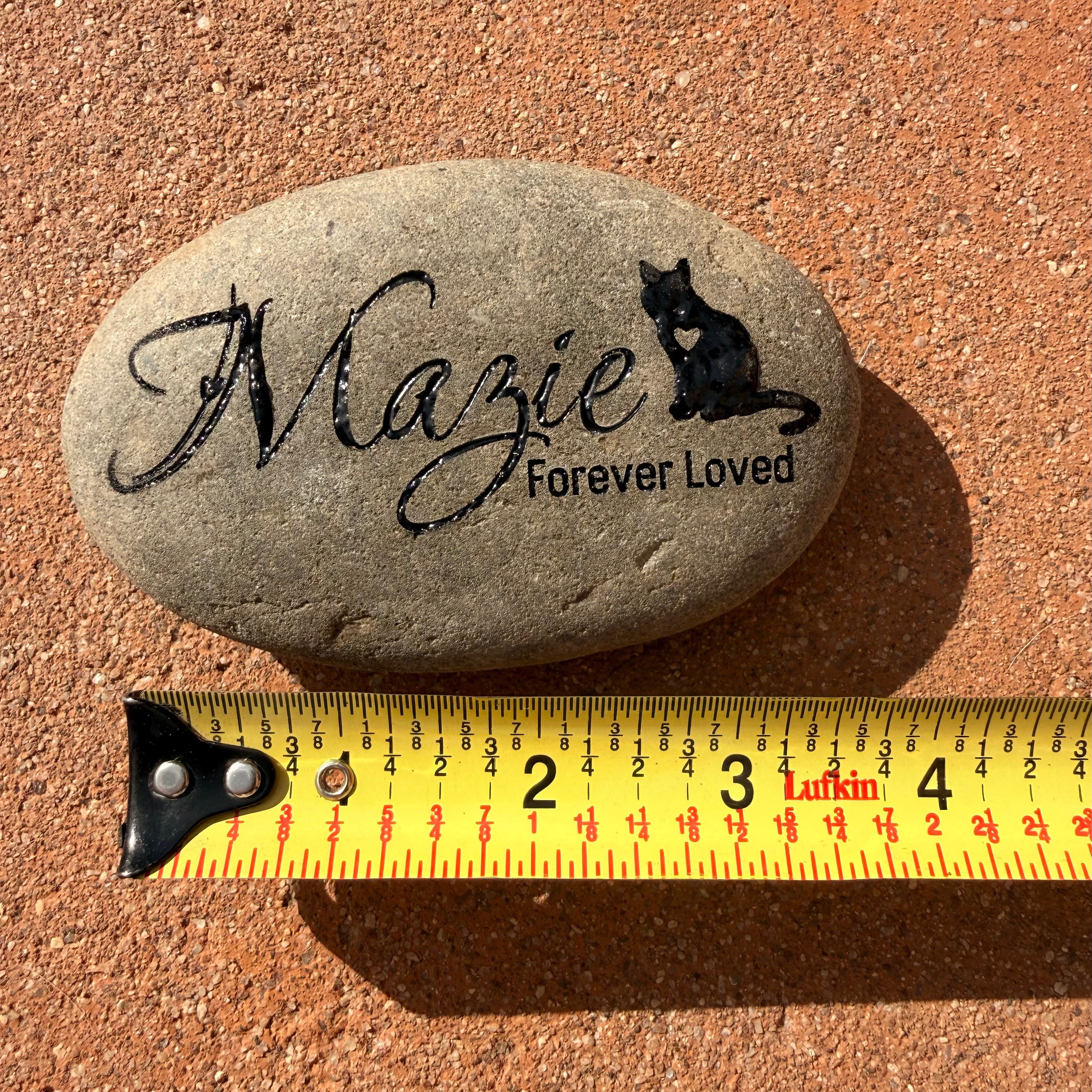 Decorative stone with 'Mazie Forever Loved' engraving and a black cat silhouette, measured against a ruler on a brown surface.