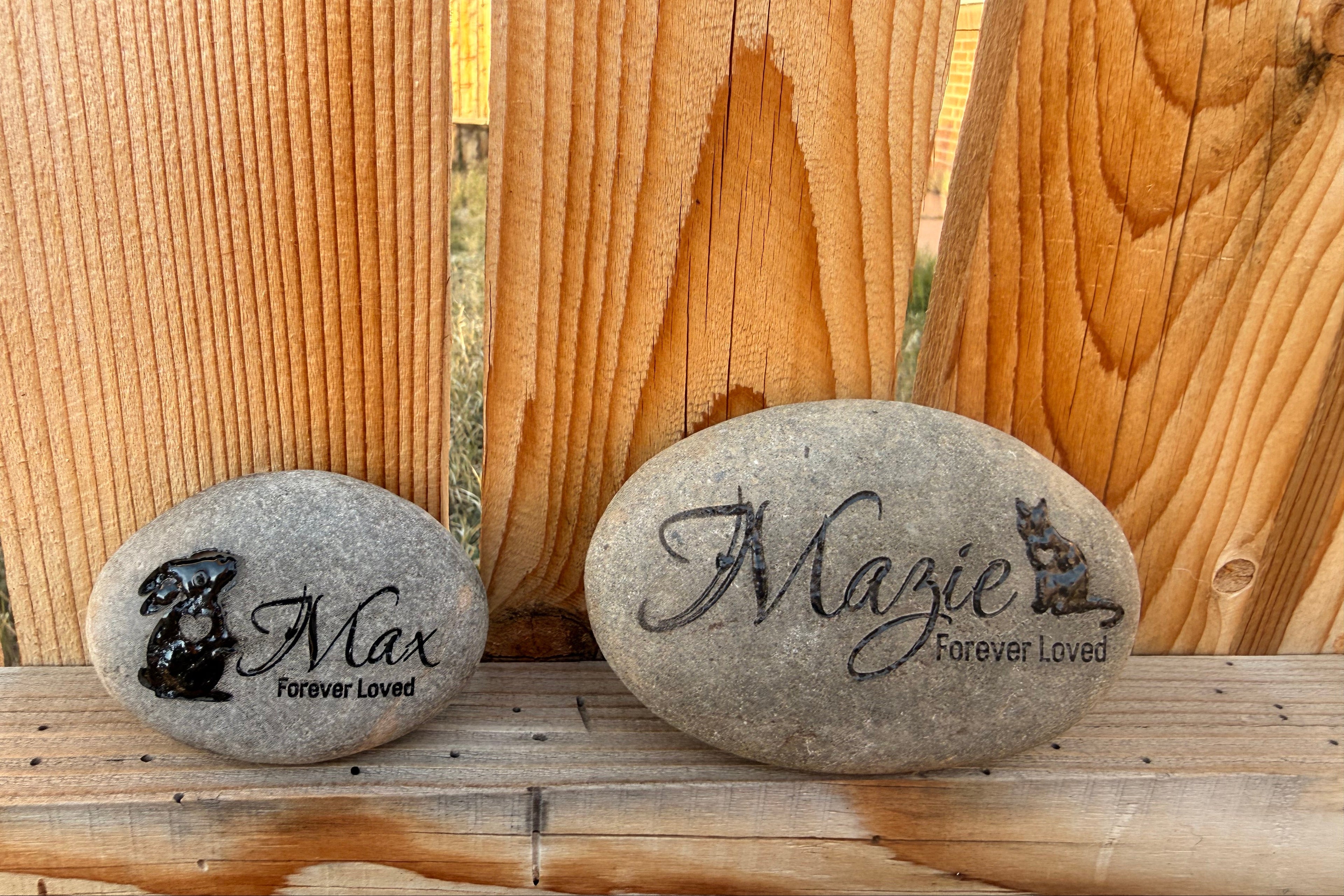 Two engraved pet memorial stones with names and animal illustrations against a wooden fence to illustrate size (3 inches-5 inches).